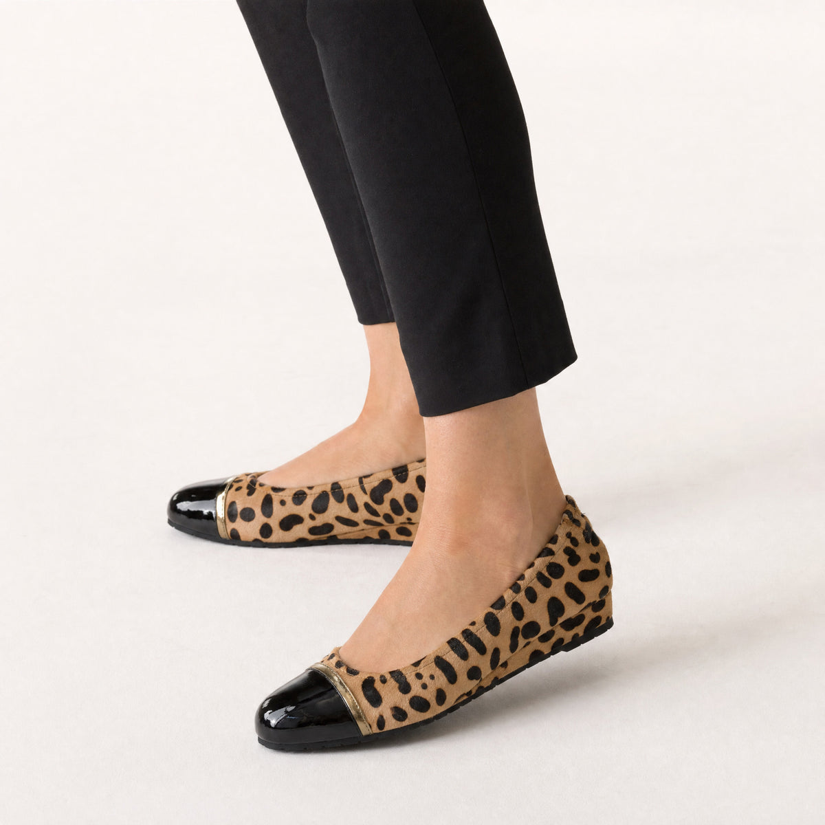 Woman standing in Journey Cleo leopard cap-toe flats with black patent toes and cropped black trousers, worn on foot